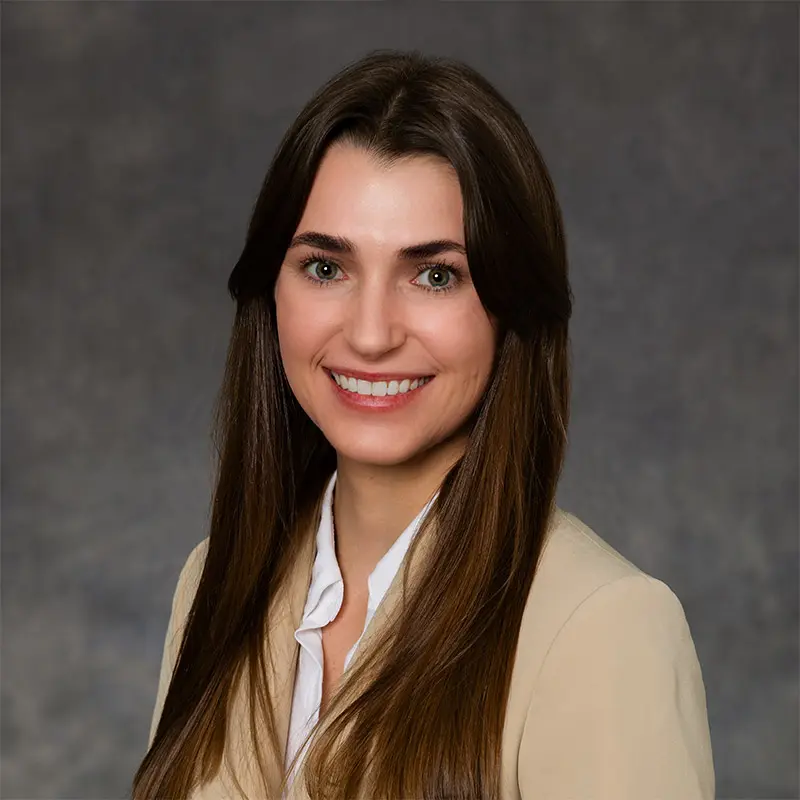 A woman with long brown hair, wearing a beige blazer and white shirt, smiles at the camera in front of a gray background.