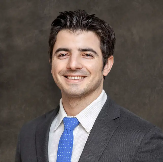 A man in a dark suit, white shirt, and blue tie smiles in front of a plain dark background.