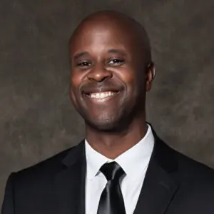 A man in a black suit and tie smiles at the camera against a dark, textured background.