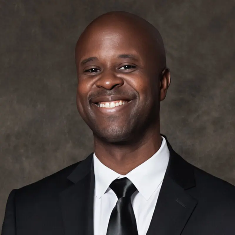A man in a black suit and tie smiles at the camera against a dark, textured background.