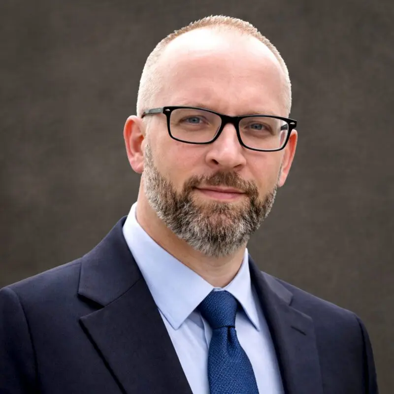 A man with a short beard and glasses wearing a dark suit, light blue shirt, and blue tie, posed against a plain dark background.