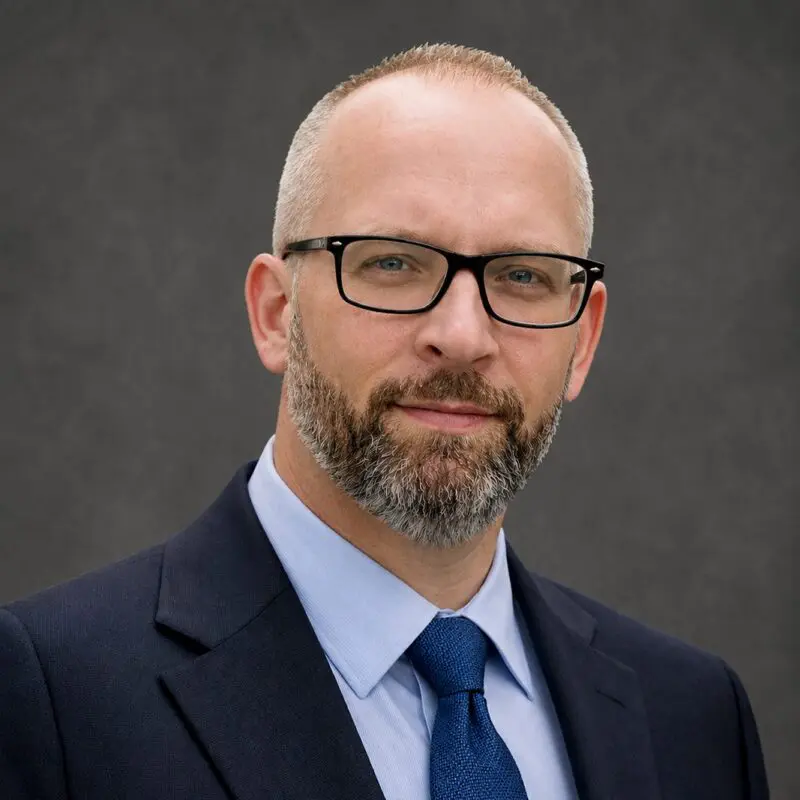 A man with glasses and a short beard wearing a dark suit, light blue shirt, and blue tie, posed against a plain dark background.