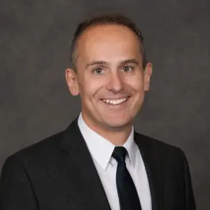 A man in a dark suit, white shirt, and black tie smiles at the camera against a plain dark gray background.