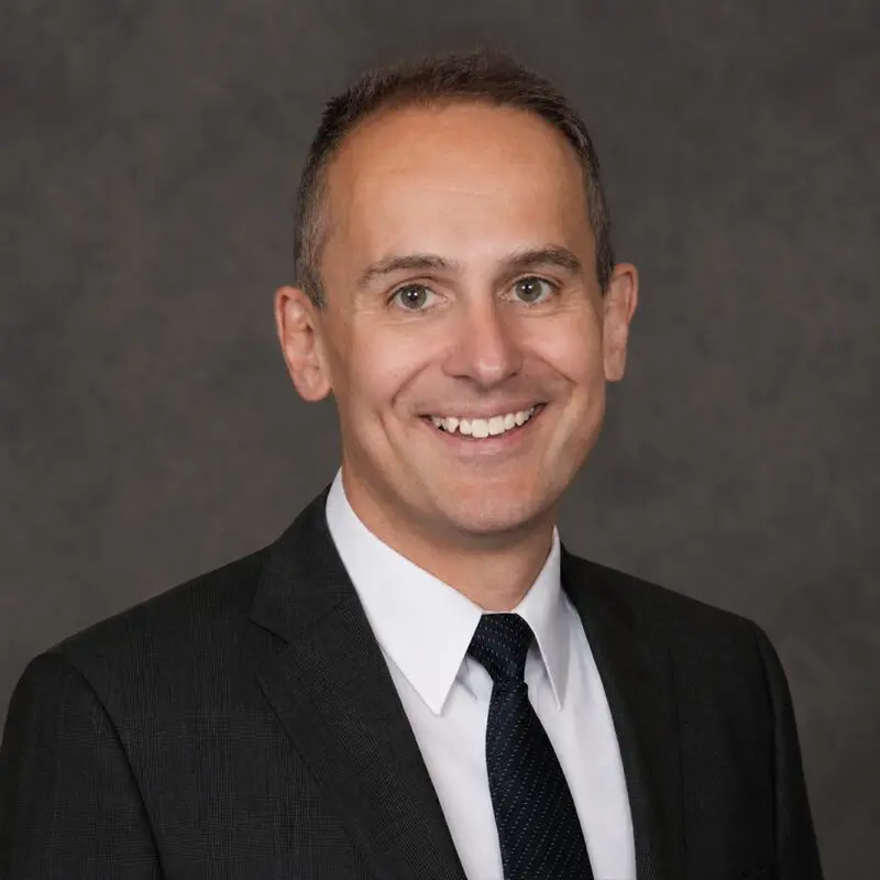 A man in a dark suit, white shirt, and black tie smiles at the camera against a plain dark gray background.