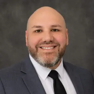 A bald man with a beard, wearing a gray suit, white shirt, and black tie, smiling in front of a plain dark gray background.