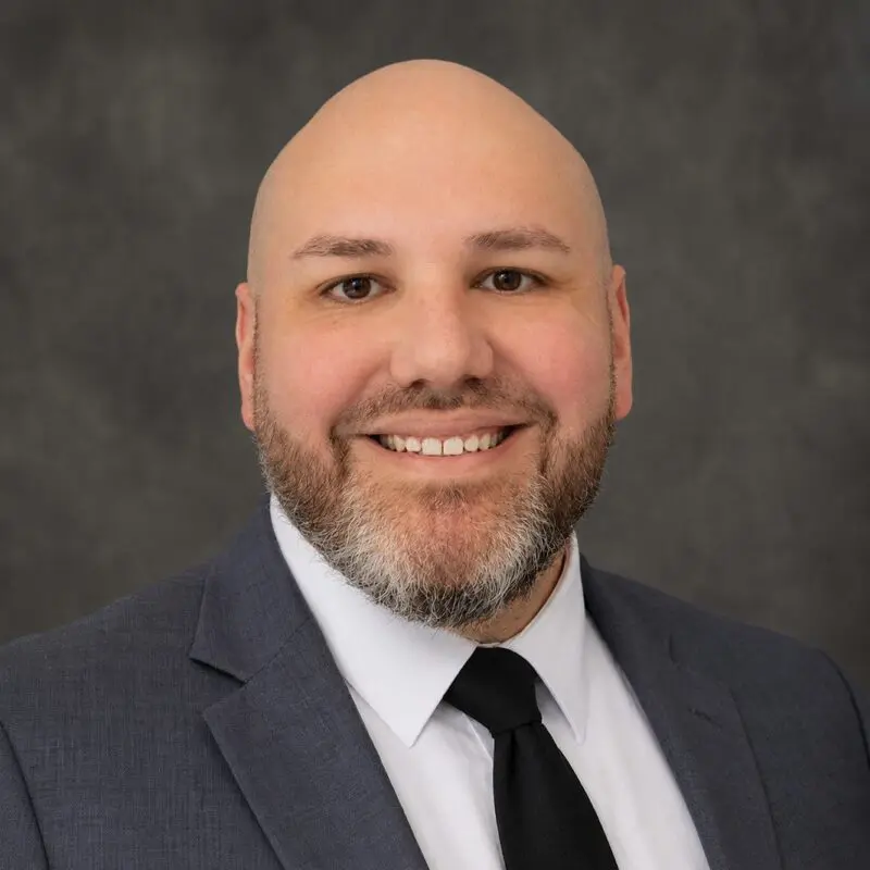 A bald man with a beard, wearing a gray suit, white shirt, and black tie, smiling in front of a plain dark gray background.