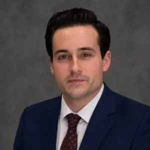 A man with short dark hair wearing a dark blue suit, white shirt, and red patterned tie poses in front of a gray background.