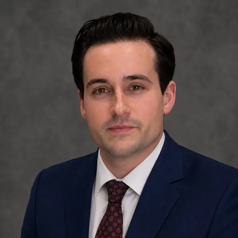 A man with short dark hair wearing a dark blue suit, white shirt, and red patterned tie poses in front of a gray background.