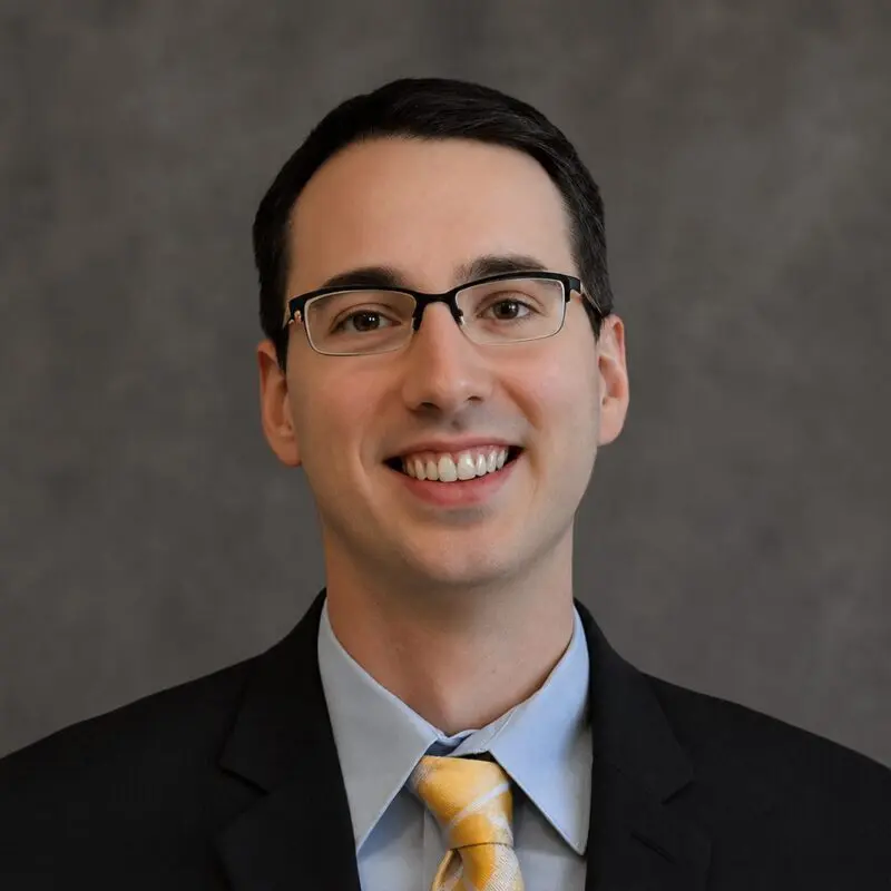 A man wearing glasses, a black suit, a light blue shirt, and a yellow tie, smiling in front of a plain gray background.