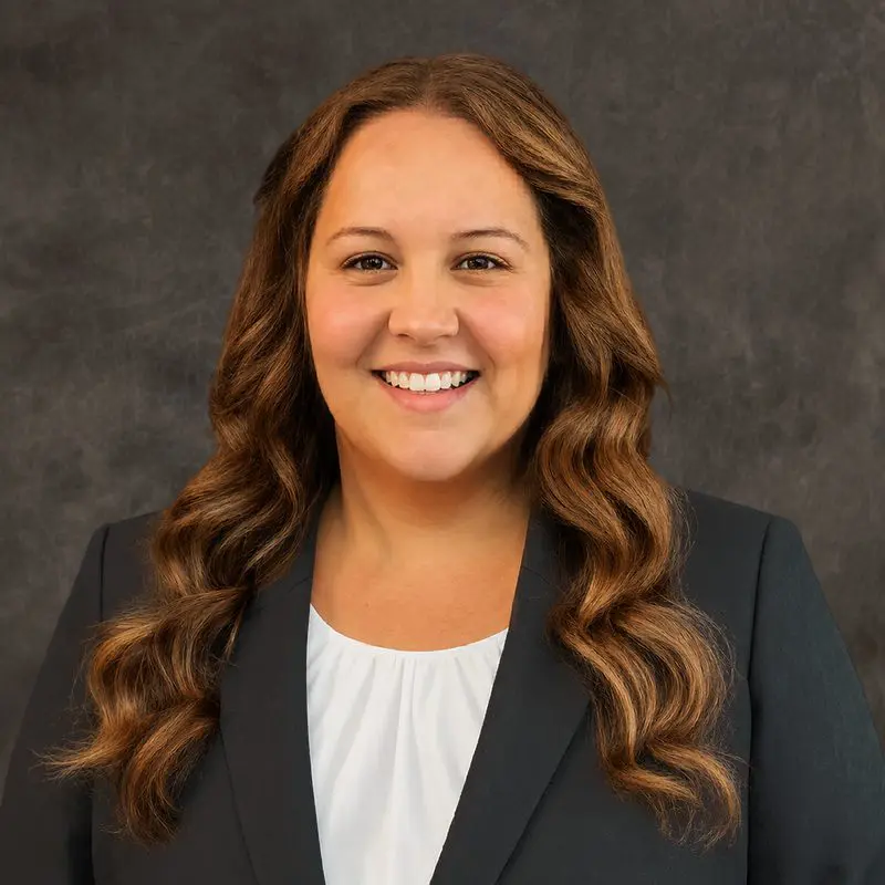 Woman with long wavy brown hair wearing a dark blazer and white blouse, smiling in front of a dark textured background.