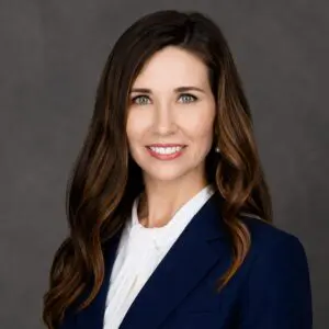Woman with long brown hair wearing a navy blazer and white blouse, smiling at the camera against a plain dark background.