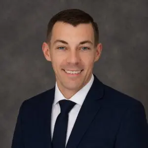 A man in a dark suit, white shirt, and dark tie poses for a professional headshot against a gray background.