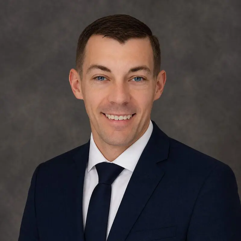 A man in a dark suit, white shirt, and dark tie poses for a professional headshot against a gray background.