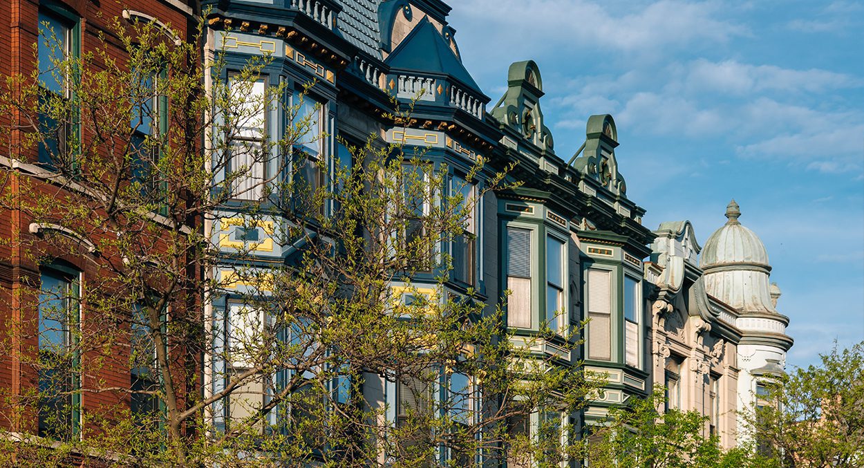 Row of historic, ornate townhouses with bay windows and decorative rooflines, partially obscured by leafy trees under a blue sky.