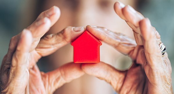 A person holds a small red house-shaped object between their hands, which are positioned to form a protective gesture around it.