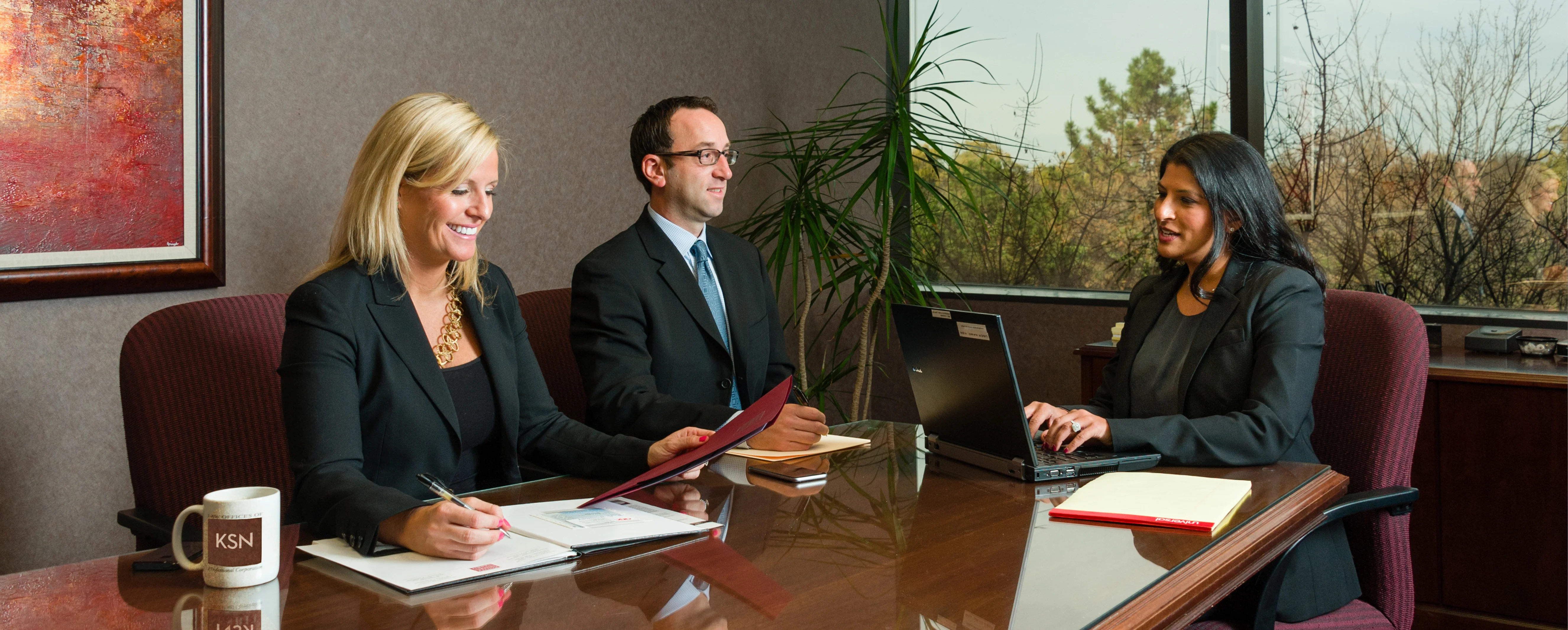 Three professionals in business attire sit at a conference table with notebooks and a laptop, engaged in a meeting in a modern office with large windows.