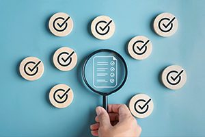 A hand holds a magnifying glass over a checklist icon, surrounded by wooden circles with check marks on a blue background.