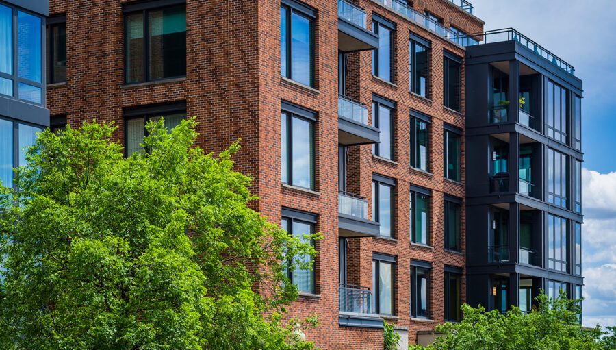 Modern brick apartment building with large windows and balconies, partially obscured by green trees, under a partly cloudy sky.