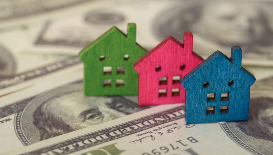 Three small wooden house models in green, red, and blue are placed on top of several US $100 bills.