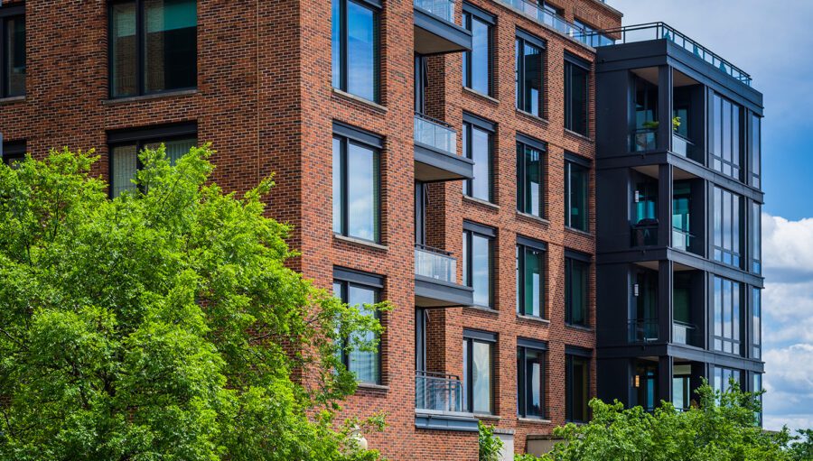 Modern brick apartment building with large windows and glass balconies, partially obscured by leafy green trees under a blue sky with clouds.