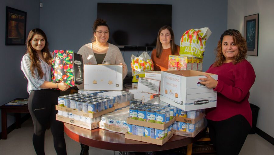 Four women stand around a table filled with canned goods and boxed food items, holding various food packages in an office setting.