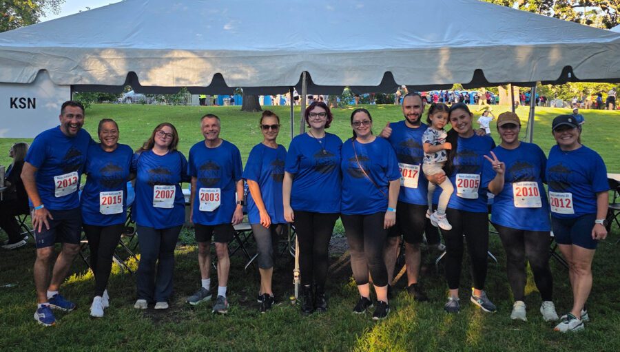 A group of eleven people, all wearing blue shirts and numbered race bibs, pose together in front of a tent at an outdoor event. One person is holding a child.