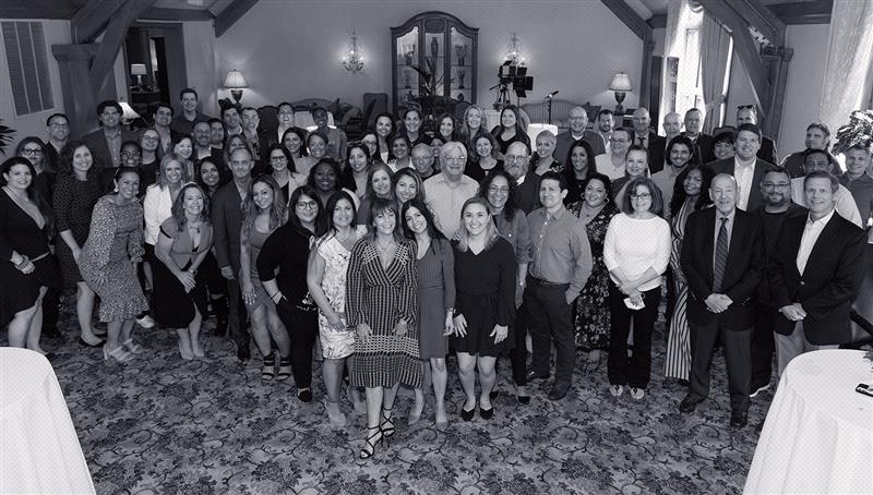 A large group of professionally dressed people pose together for a group photo in a well-lit room with ornate decor and carpet.