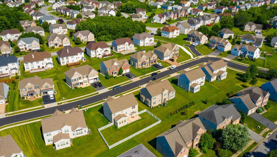 Aerial view of a suburban neighborhood with rows of single-family houses, green lawns, trees, and winding roads on a sunny day.