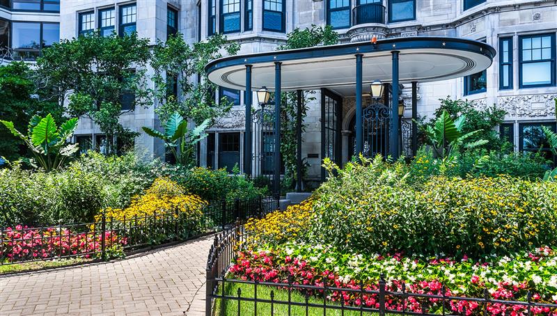 A landscaped garden with colorful flowers surrounds a paved walkway leading to the entrance of a stone building with black columns and decorative details.