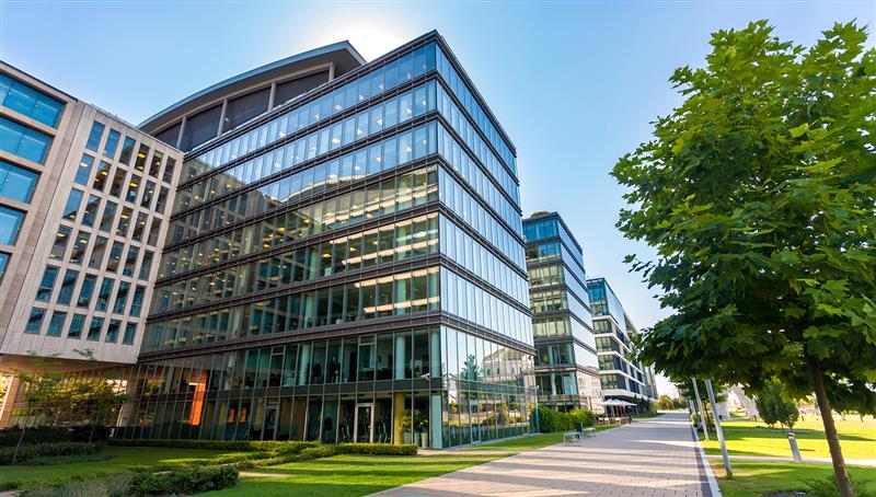 Modern glass office buildings with reflective windows, located alongside a landscaped walkway and green trees on a sunny day.