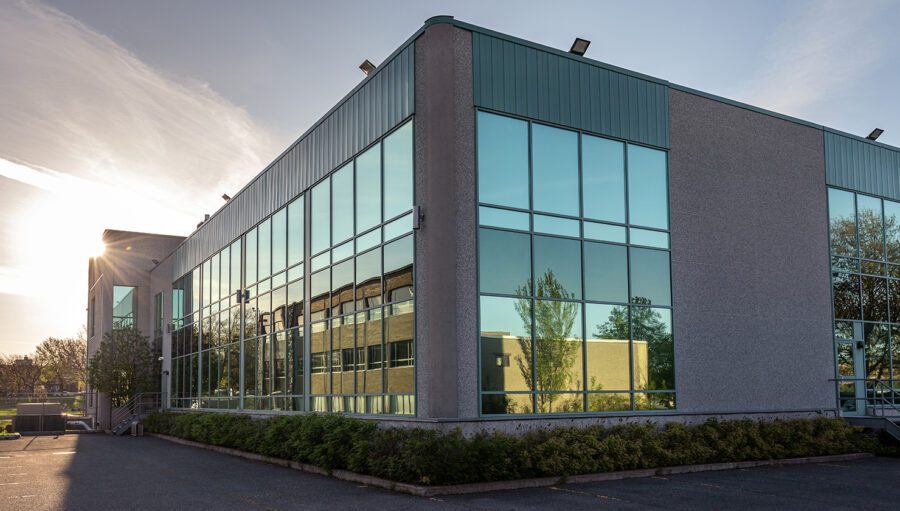 Modern commercial building with large reflective glass windows, concrete walls, and a green roof trim, photographed at sunset with trees and another building visible in the reflection.