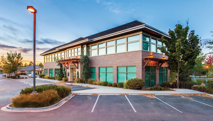 Modern two-story office building with large windows, brick exterior, and a nearly empty parking lot at sunset.