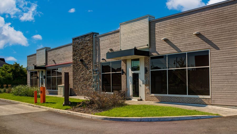 A modern, empty commercial building with large glass windows and stone accents, set on a sunny day with a neatly maintained lawn and a paved driveway.
