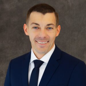 A man in a navy suit and tie smiles at the camera against a plain dark gray background.